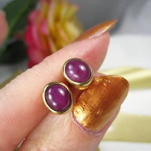 May include: A pair of oval stud earrings with a gold-coloured setting. Each earring features a deep purple, polished stone. The earrings are displayed against a soft background, highlighting their colour and design.