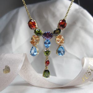 May include: A gold necklace with a multi-coloured gemstone pendant. The pendant features a variety of gemstones, including red, orange, yellow, green, blue, and purple. The necklace is on a white background.