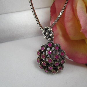 May include: A silver-toned pendant necklace featuring a floral design with small, round, pink gemstones. The pendant hangs from a delicate chain. The necklace is displayed against a white background with a pink and yellow rose.