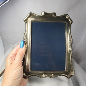 May include: A rectangular silver-toned metal picture frame with a polished finish and decorative, curved edges. The frame features a dark navy blue velvet backing insert, providing a deep contrast to the metallic border of the frame.