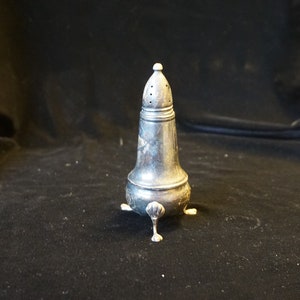 May include: A silver salt shaker with a conical top and small dispensing holes. The antique shaker has a bulbous base and rests on three small, decorative feet. The design is ornate and vintage, typical of older tableware.