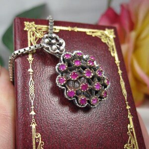May include: A silver pendant necklace with a cluster of small, round, pink gemstones. The pendant is set in a silver filigree design and hangs on a delicate silver chain. The necklace is displayed on a burgundy surface with gold accents.