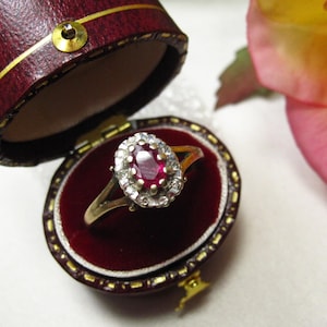 Könnte beinhalten: Ein goldener Ring mit einem rubinroten Mittelstein, umgeben von kleinen weißen Steinen, präsentiert in einer offenen, bordeauxfarbenen Schmuckschatulle. Die Schatulle hat einen goldenen Rand und ein rotes Samtinterieur. Eine Rose ist im Hintergrund.