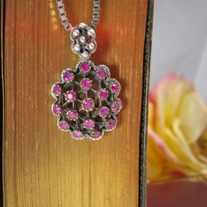 May include: A silver pendant necklace with a floral design. The pendant features a cluster of small, round, pink gemstones set in a silver frame. The necklace is displayed against a backdrop of a vintage book and a blurred yellow flower.