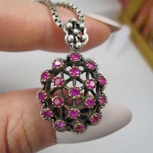 May include: A silver pendant necklace with a floral design. The pendant features a cluster of small, round, bright pink gemstones set within a silver filigree frame. The necklace chain is a delicate silver box chain.