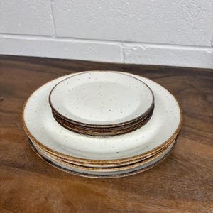 Pode incluir: Um conjunto de pratos de cerâmica branco-sujo com um design salpicado e uma borda marrom. A pilha inclui pratos grandes e pratos menores, todos com uma estética rústica. Os pratos estão dispostos sobre uma superfície de madeira.