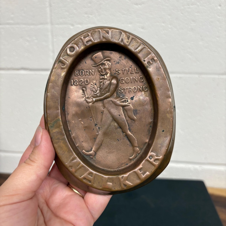 Op de afbeelding: Ovale, koperkleurige metalen plaat met een wandelende figuur in hoge hoed en jas, die een wandelstok vasthoudt. De tekst "JOHNIE WALKER" en "BORN 1820 STILL GOING STRONG" zijn in reli&euml;f. De plaat wordt door een hand vastgehouden.