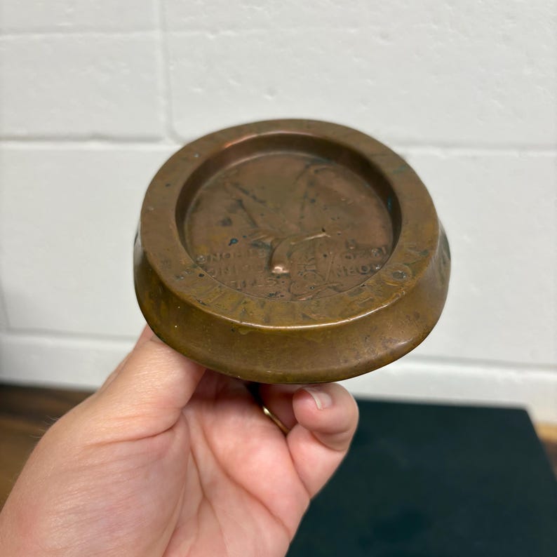 Op de afbeelding: Een rond, bronskleurig object met een opstaande rand en een gedetailleerd centraal ontwerp. Het ontwerp lijkt een reli&euml;f van een persoon en tekst te zijn. Het object wordt in een hand gehouden.