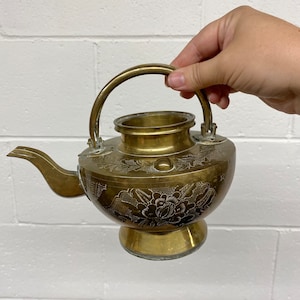 May include: A brass teapot with an ornate floral design. The teapot has a round body and a curved spout. The handle is attached to the top of the teapot and has a decorative design.