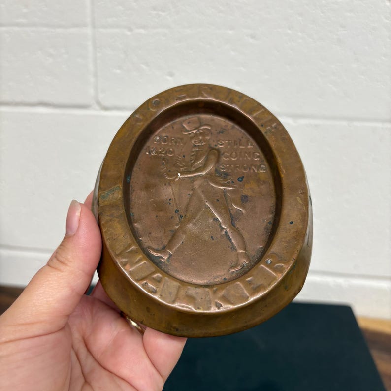 Op de afbeelding: Een bronzen asbak met een cirkelvormig middenstuk met een reli&euml;f van een man die loopt. De tekst "BORN 1820 STILL GOING STRONG" en "WALKER" zijn zichtbaar. De asbak heeft een afgeronde, onregelmatige vorm.