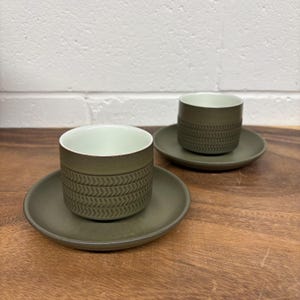 May include: Two green ceramic coffee cups with matching saucers. The cups have a white interior and a textured, geometric pattern on the exterior.