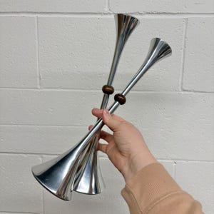 May include: Two silver-colored metal vases with a flared trumpet shape. Each vase has a slender stem and a dark brown wooden bead accent. The vases are held against a white brick wall.
