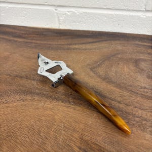 May include: A vintage bottle opener with a chrome head and a brown, marbled handle. The opener is designed for removing bottle caps and features a classic design. The handle is approximately 13 cm long.