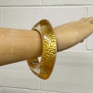 May include: A clear, gold-toned bangle bracelet with a textured, hammered metal appearance. The bracelet is thick and rounded, with a transparent quality. The jewelry is displayed on a wooden arm.