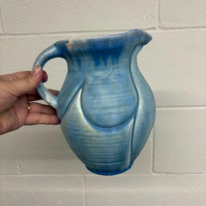 Puede incluir: Una jarra de cerámica azul con asa y pico vertedor. La jarra tiene una superficie texturizada y una forma bulbosa con ranuras verticales. El color pasa de azul claro a azul más oscuro en la parte superior e inferior.