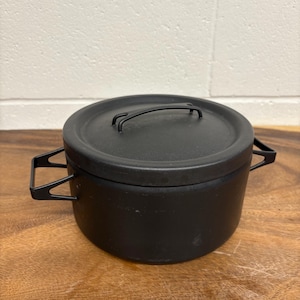 Op de afbeelding: Een zwarte gietijzeren pot met een deksel en twee handvatten aan de zijkant. De pot is rond en heeft een matte afwerking. Het deksel heeft een handvat om gemakkelijk op te tillen. De pot staat op een houten oppervlak.