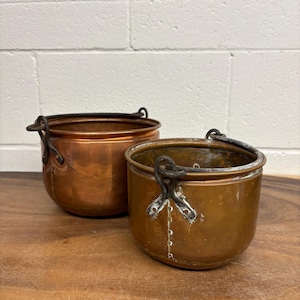 Peut inclure: Deux pots en cuivre antiques avec des poignées en métal foncé. Les pots ont une forme arrondie et une couleur brun-rougeâtre chaude. Ils sont posés sur une surface en bois devant un mur de briques blanches.