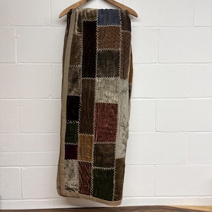 Op de afbeelding: Een patchwork plaid met een verscheidenheid aan fluwelen vierkanten in aardetinten. De deken hangt aan een houten hanger tegen een witte bakstenen muur. De vierkanten zijn in tinten bruin, groen en bordeaux, met een beige rand.
