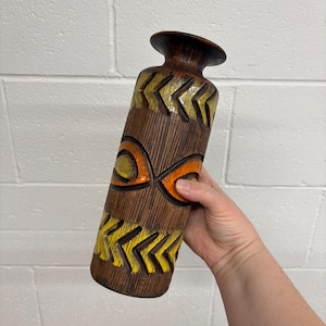 May include: A brown ceramic vase with a textured surface and a cylindrical shape. The vase features yellow and orange geometric designs. The vase is approximately 12 inches tall.
