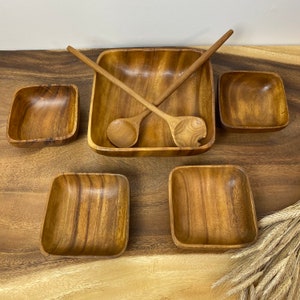 May include: A set of five wooden bowls and two wooden spoons. The bowls are square and rectangular, and the spoons are long and thin. The bowls are all made of the same type of wood, which has a light brown colour.
