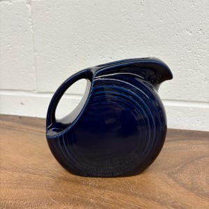 May include: A navy blue ceramic pitcher with a distinctive handle and spout. The pitcher has a rounded body with a series of concentric ridges. The handle is a large, open loop. The pitcher is sitting on a wooden surface.
