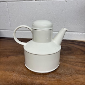 May include: A white ceramic teapot with a rounded body, a curved handle, and a spout. The teapot has a lid with a small knob and sits on a wooden surface. The background is a white brick wall.