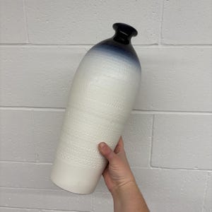 May include: A tall, ceramic vase with a textured, off-white body and a gradient of blue to white. The vase has a narrow, dark blue neck and a wide base. The body features a repeating pattern of small circles.