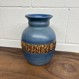 Può includere: Un vaso di ceramica blu con una fascia strutturata a forma di corteccia al centro. Il vaso ha un corpo arrotondato e un bordo leggermente svasato. La superficie ha un aspetto maculato e poggia su una superficie di legno.
