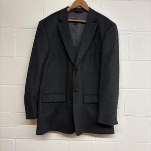 Puede incluir: Un blazer gris oscuro con cierre de dos botones, dos bolsillos delanteros y tres botones en cada manga. El blazer está colgado en una percha de madera contra una pared de ladrillos blancos.