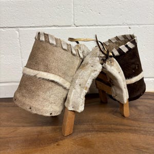 May include: A pair of footstools featuring a cowhide exterior in shades of light beige and dark brown. Each stool is supported by four wooden legs and includes a decorative whip-stitched trim along the top edge and a horizontal band of white fur.