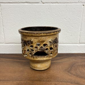 May include: A ceramic planter with a textured, beige exterior and a dark brown interior. The planter features decorative cutouts around the middle and sits on a short, pedestal base. The interior is visible through the cutouts.