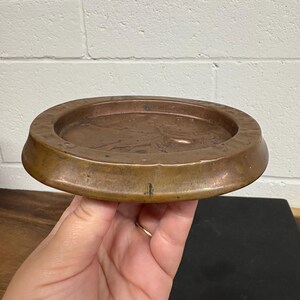 Op de afbeelding: Een ronde, antieke bronzen asbak met een opstaande rand en een ondiep bekken. De asbak heeft een donkere, verouderde patina, wat suggereert dat hij vintage is. Het ontwerp is eenvoudig en functioneel, met een klassieke esthetiek.