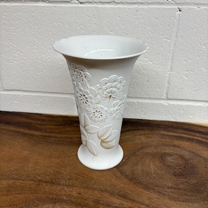 May include: White ceramic vase with embossed floral and leaf designs. The vase has a flared top and a pedestal base. The floral patterns are detailed and create a textured surface. The vase is displayed on a wooden surface.