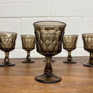Könnte beinhalten: Set aus sechs braun getönten Vintage-Stielgläsern. Jedes Glas hat ein rautenförmiges Muster auf der Schale und einen zarten Stiel. Die Gläser sind auf einer Holzoberfläche angeordnet, mit einer weißen Backsteinmauer im Hintergrund.