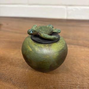 May include: A small, round, green jar with a turtle figurine on top. The turtle is green with a blue eye. The jar has a dark green and gold finish. The turtle is resting on a black ring.