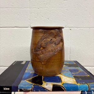 Può includere: Un vaso in ceramica marrone con una superficie strutturata e un disegno intagliato di un pesce. Il vaso ha una forma cilindrica con un bordo leggermente svasato. Il vaso è appoggiato su un libro.