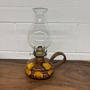 May include: A vintage oil lamp with a clear glass globe and a brown ceramic base. The base features a pattern of yellow and brown spots, and it has a curved handle. The lamp is designed to sit on a table.