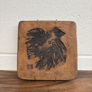 Puede incluir: Un adorno de pared cuadrado de cerámica marrón con un relieve de un pájaro. El pájaro tiene plumas detalladas y un tocado decorativo. Dos bucles metálicos están unidos en la parte superior para colgarlo.