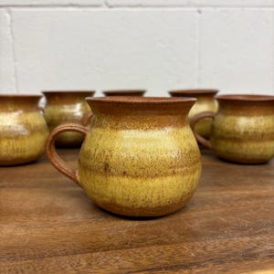 May include: A collection of rounded ceramic mugs featuring a two-tone glaze with a warm yellow centre and dark brown rims and bases. Each mug has a curved handle and a wide, circular opening, showcasing a smooth, glossy finish throughout.
