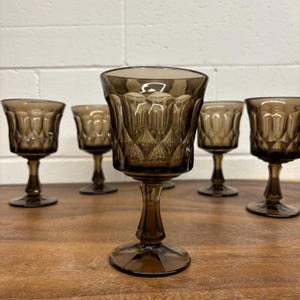 Puede incluir: Juego de seis copas de vino teñidas de marrón. Cada copa presenta un patrón decorativo texturizado en el cuenco y una base con tallo. Las copas están dispuestas sobre una superficie de madera.