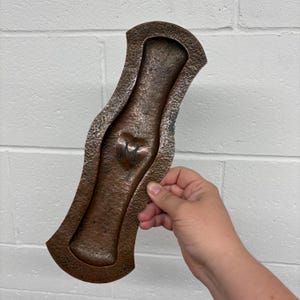 May include: An elongated, hammered copper tray with a curved, hourglass-like silhouette. The surface features a textured, dimpled finish and a central, embossed heart shape containing the letter M. The metal has a dark, aged patina throughout.