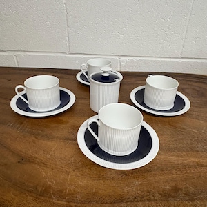 May include: A white porcelain tea set with a ribbed design. The set includes four teacups, saucers, and a sugar bowl with a lid. The saucers and the sugar bowl have a dark blue centre with a white border. The teacups have handles.