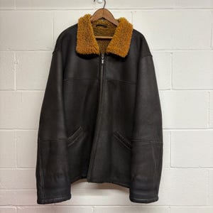 May include: A brown leather jacket with a tan shearling collar and lining. The jacket has a zip-up front, two front pockets, and is hanging on a wooden hanger. The jacket appears to be vintage.