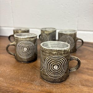May include: Set of six ceramic mugs with a dark brown, textured finish. Each mug features a white concentric circle design. The mugs have a handle and are displayed on a wooden surface.