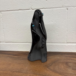May include: A black ceramic sculpture of a draped figure with turquoise accents and a floral pattern. The sculpture stands on a wooden surface against a white brick wall. The figure is a stylized representation.