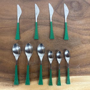 Puede incluir: Un juego de cubiertos vintage con hojas y cucharas de acero inoxidable y mangos de plástico verde. El juego incluye cuchillos y cucharas de varios tamaños, dispuestos sobre una superficie de madera.