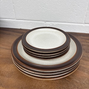 May include: A set of ceramic plates featuring a speckled beige centre and a dark brown border. The plates are stacked on a larger plate with the same design. The plates are on a wooden surface.