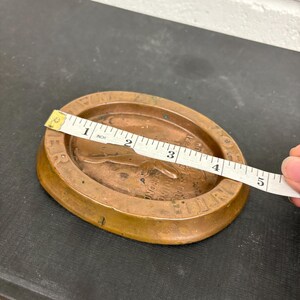 Op de afbeelding: Een bronzen ovale schaal met een opstaande rand. De schaal heeft een reli&euml;f ontwerp met tekst en een figuur. Een meetlint toont aan dat de schaal ongeveer 10 cm lang is.