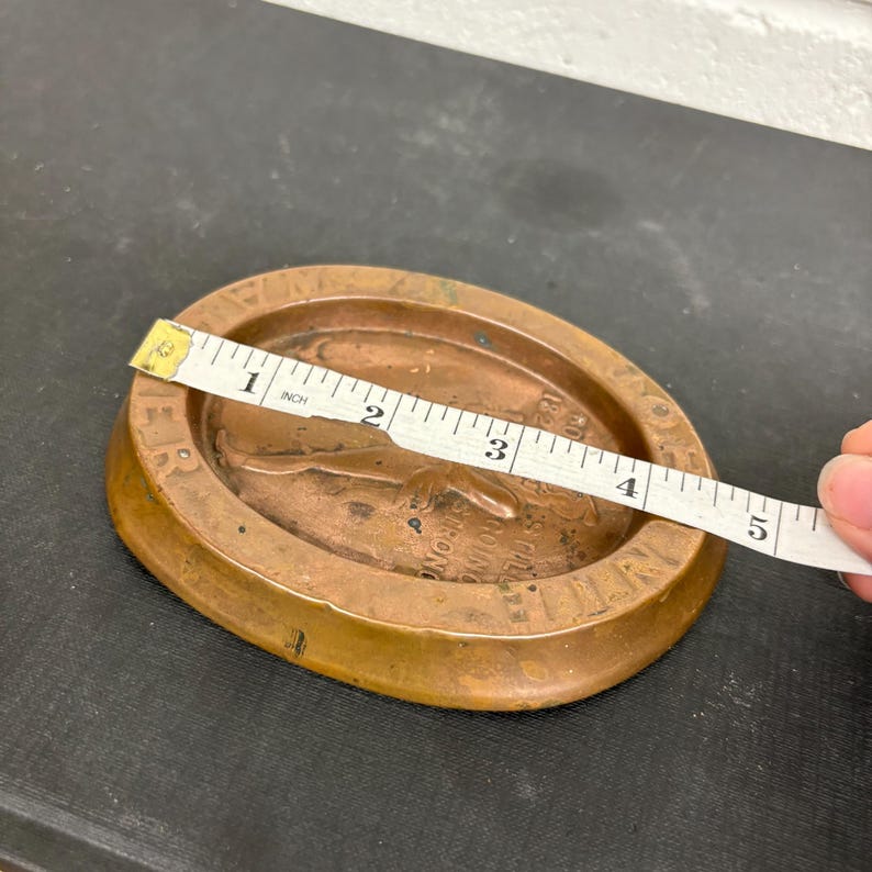 Op de afbeelding: Een bronzen ovale schaal met een opstaande rand. De schaal heeft een reli&euml;f ontwerp met tekst en een figuur. Een meetlint toont aan dat de schaal ongeveer 10 cm lang is.