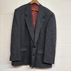 May include: A dark gray blazer with a two-button closure, two front pockets, and a notched lapel. The interior lining is a deep red color. The blazer is hanging on a wooden hanger against a white brick wall.